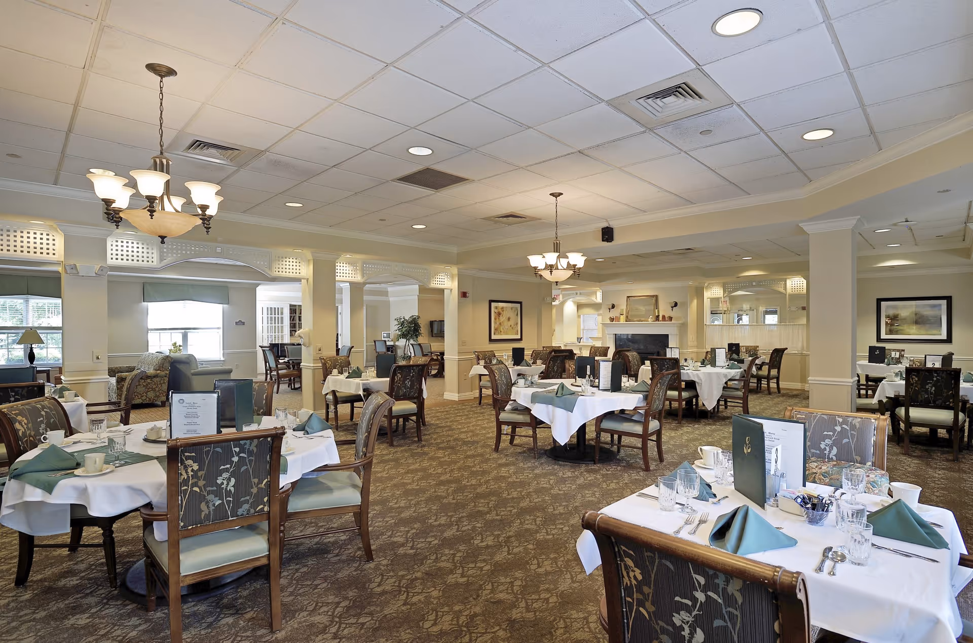 A spacious dining room with multiple round tables covered with white tablecloths and green napkins. Each table is set with glassware, cups, and menus. The room has patterned carpet, wooden chairs with floral upholstery, and warm lighting from chandeliers. There are large windows on the left side and a fireplace with a mirror above it in the background.