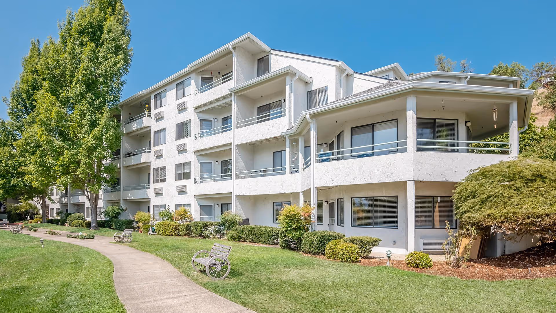 Exterior view of a multi-story white residential building with balconies, surrounded by green grass, bushes, and trees under a clear blue sky. A paved walkway curves along the front of the building with benches placed along the path.