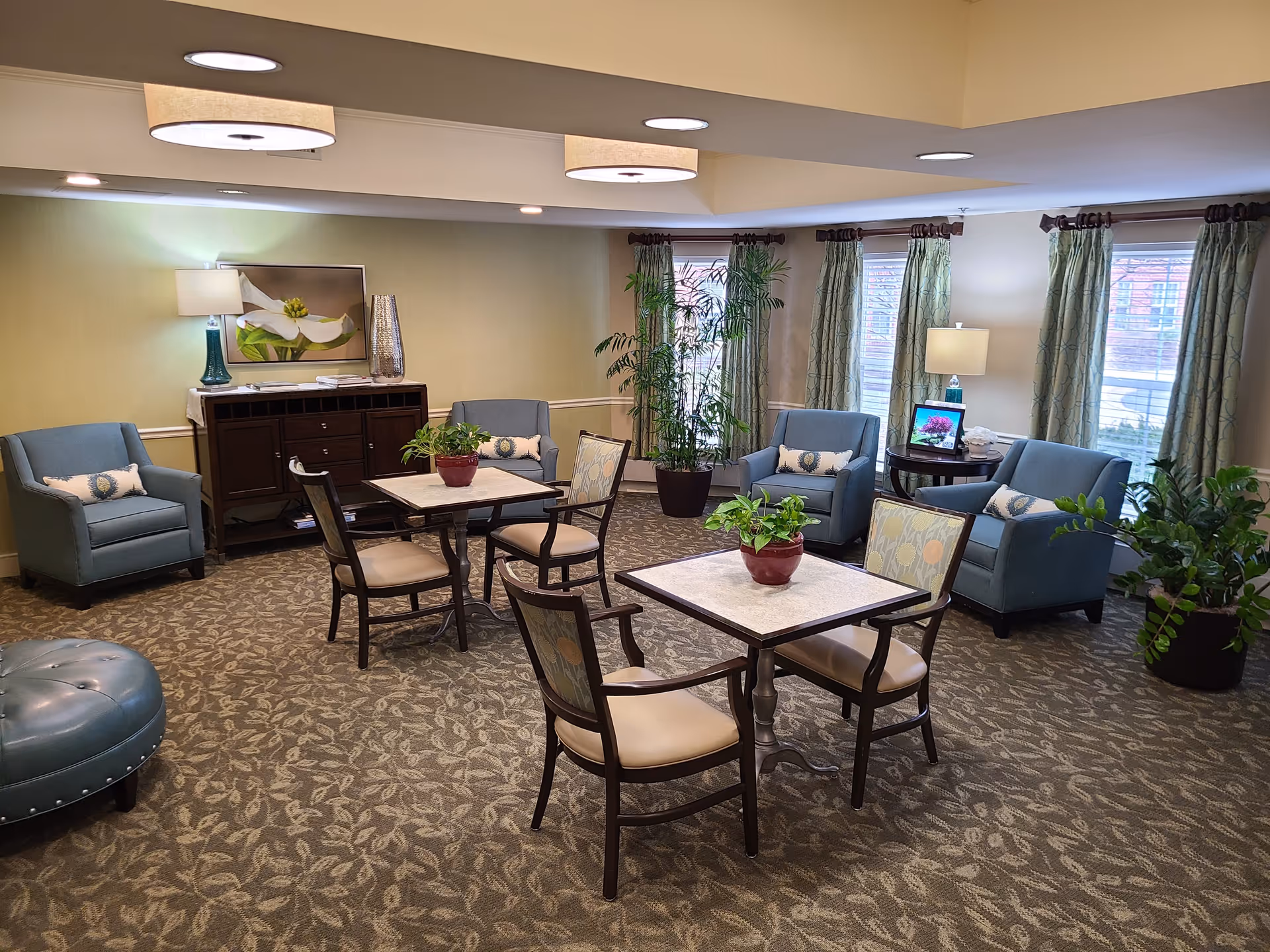 A cozy assisted living common area with two square tables each surrounded by four chairs. There are several blue armchairs and a blue loveseat arranged around the room. The room has patterned carpet, green curtains on windows, potted plants, a sideboard with a lamp and decorative items, and ceiling lights.