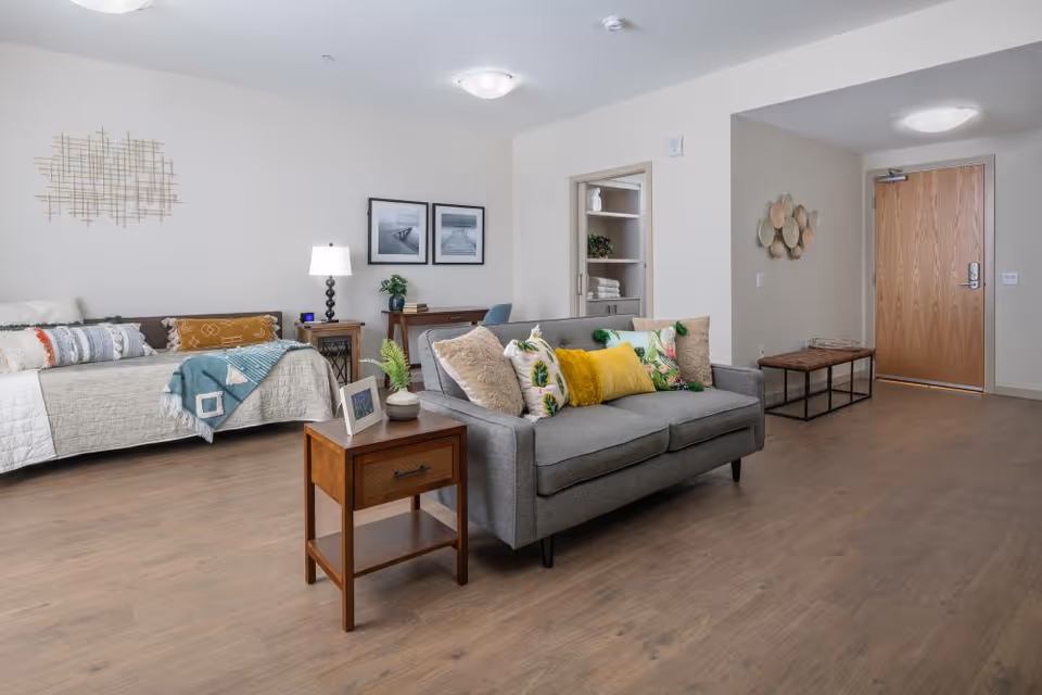 A bright and spacious senior living studio apartment featuring a bed with decorative pillows and a throw blanket, a gray sofa with colorful cushions, a wooden side table with a small plant and picture frame, a desk with a chair and lamp, and a wooden entry door. The room has light-colored walls, wood flooring, and modern wall art.