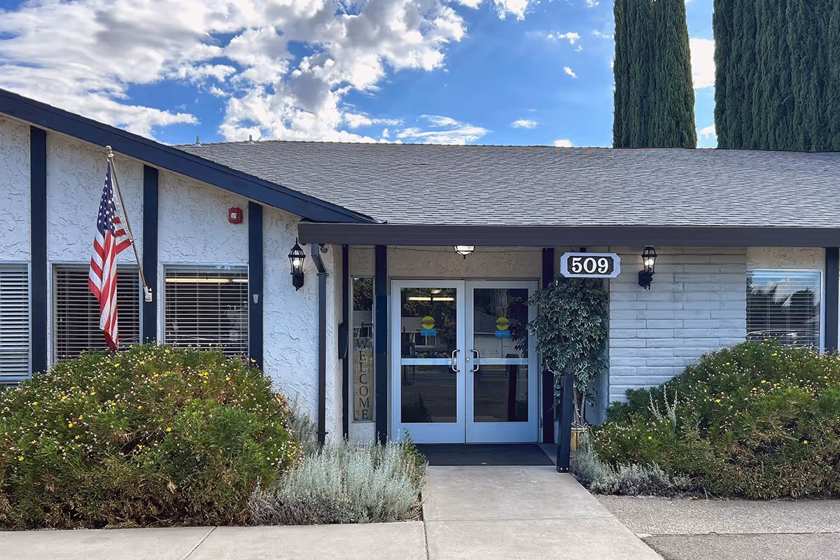 Front entrance of a single-story building with white walls and dark trim, featuring double glass doors under a small covered porch. The building number 509 is displayed above the doors. There are bushes and plants on either side of the entrance, an American flag on the left, and tall trees in the background under a partly cloudy blue sky.