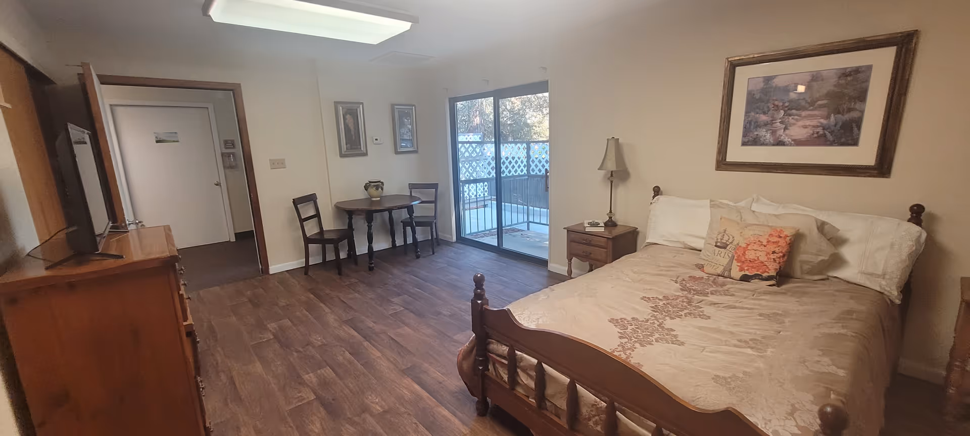 Furnished senior living studio with a bed, nightstand, dresser, small table and chairs, and a sliding glass door to a balcony.