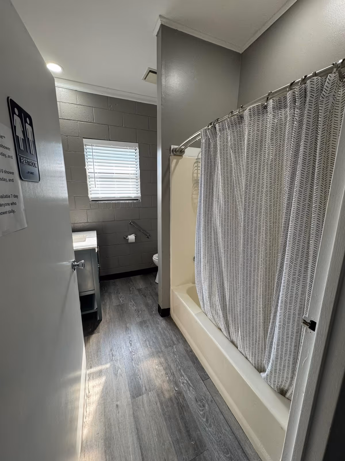 View into a restroom with a bathtub and shower curtain on the right, a window with blinds on the back wall, a toilet partially visible, and a sink with a cabinet on the left. The floor has gray wood-like vinyl planks and the walls are painted gray.