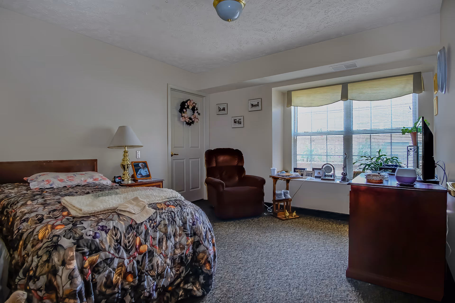 A cozy bedroom in a senior living facility featuring a single bed with a floral patterned comforter and a pillow. Next to the bed is a wooden nightstand with a brass lamp and a framed photo. A comfortable brown recliner chair is positioned near a large window with blinds and a valance. The window sill holds various decorative items including plants, clocks, and framed pictures. A wooden dresser with a TV on top is placed against the opposite wall. The room has light-colored walls, carpeted floor, and a door decorated with a floral wreath.