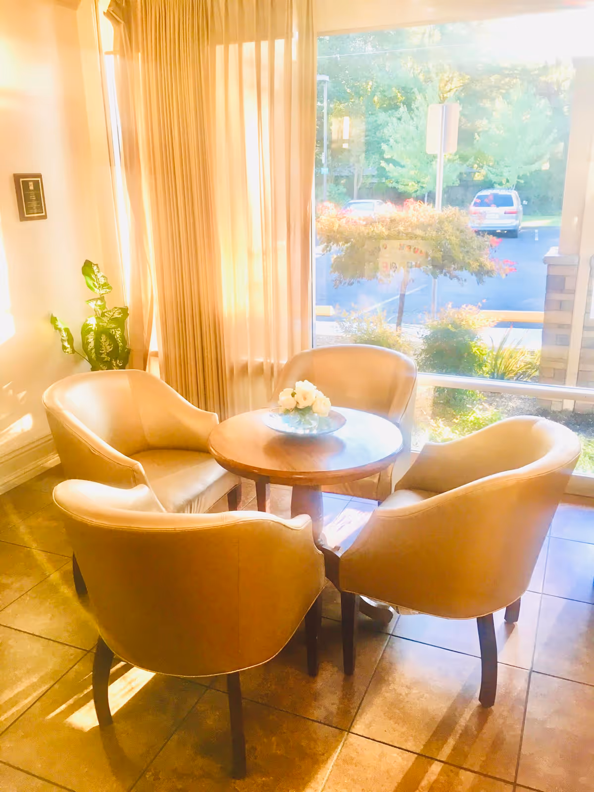 Four beige armchairs arranged around a small round wooden table with a floral centerpiece beside a sunlit window.