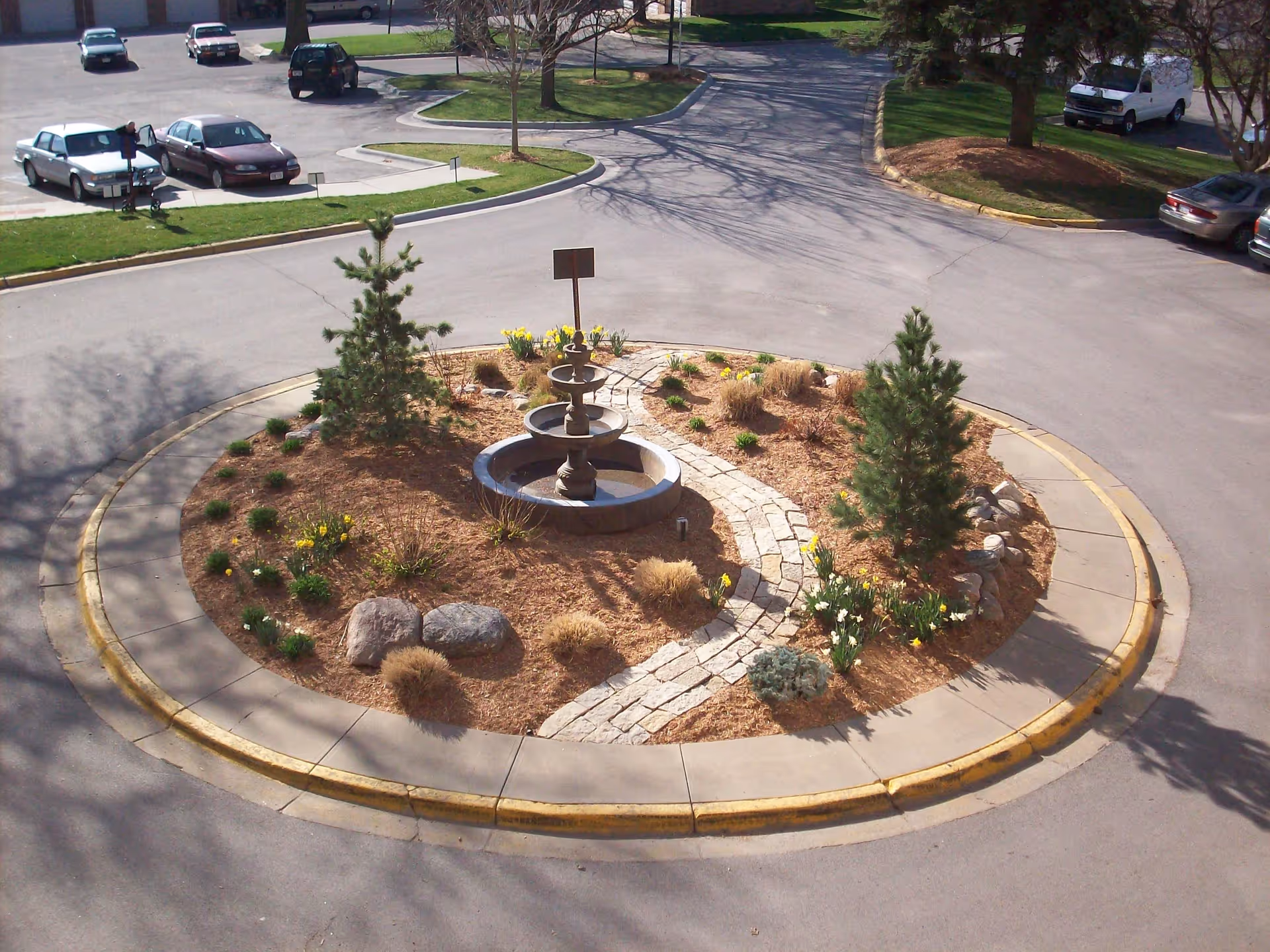 A circular landscaped island in the middle of a driveway with a stone pathway leading to a tiered water fountain surrounded by small trees, plants, and flowers. Several parked cars and a white van are visible in the background along with grassy areas and trees.
