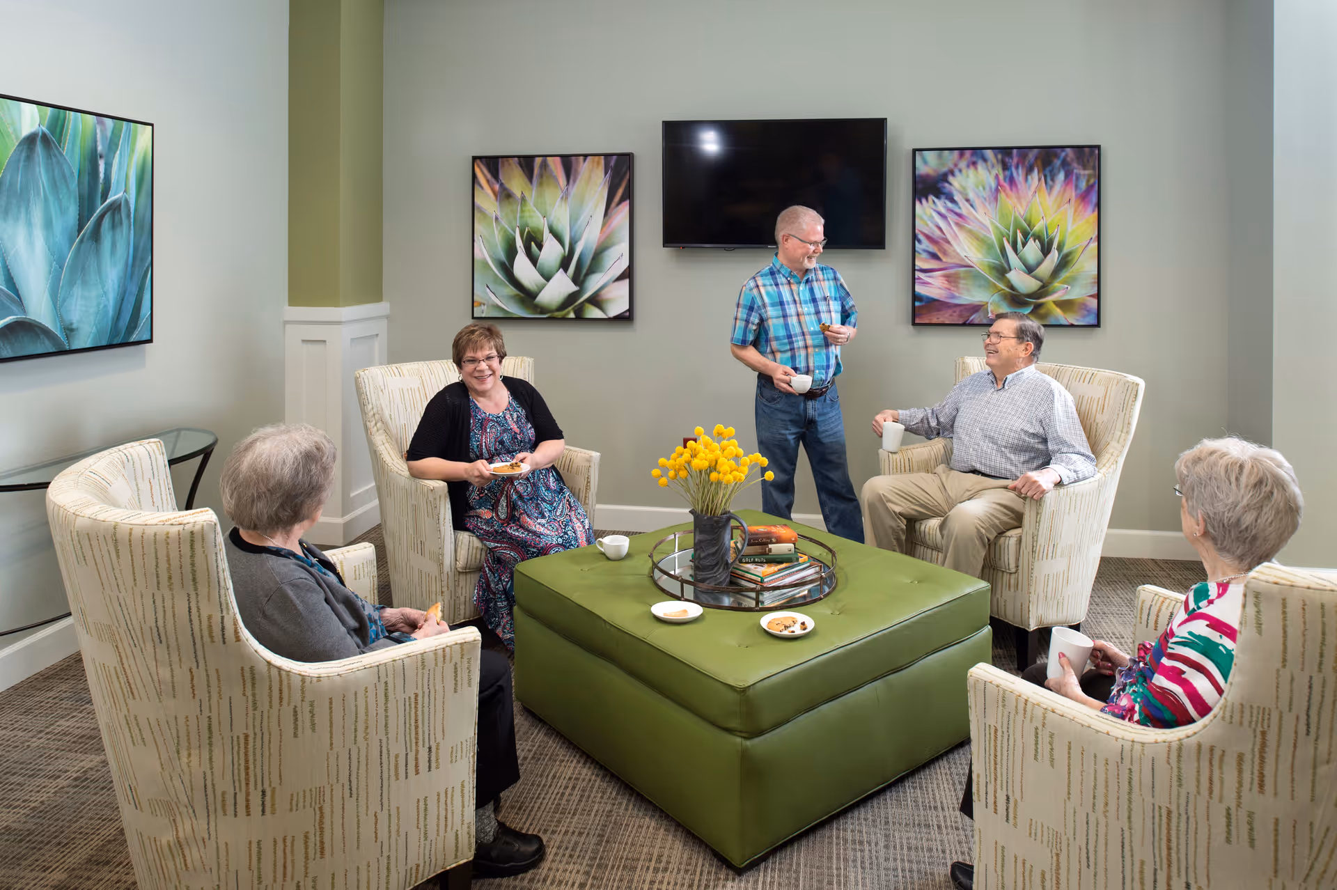A group of five elderly people socializing in a comfortable living room area with four armchairs arranged around a large green ottoman. The ottoman has a tray with books and a vase of yellow flowers. Two men and three women are holding cups and plates, enjoying snacks. The room has light green walls with three large framed pictures of succulent plants and a flat-screen TV mounted on the wall.
