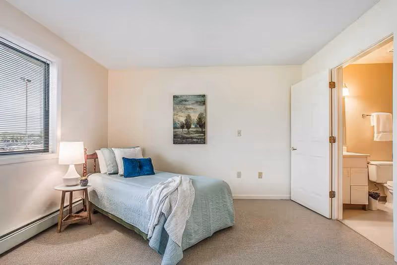 A small bedroom with a single bed covered in a light blue quilt and a white blanket draped over the foot. There are two pillows on the bed, one white and one blue. A wooden side table with a white lamp is next to the bed. A window with blinds is on the left wall, and a painting of trees hangs on the wall above the bed. An open door on the right leads to a bathroom with a toilet, sink, and towel hanging on the wall.