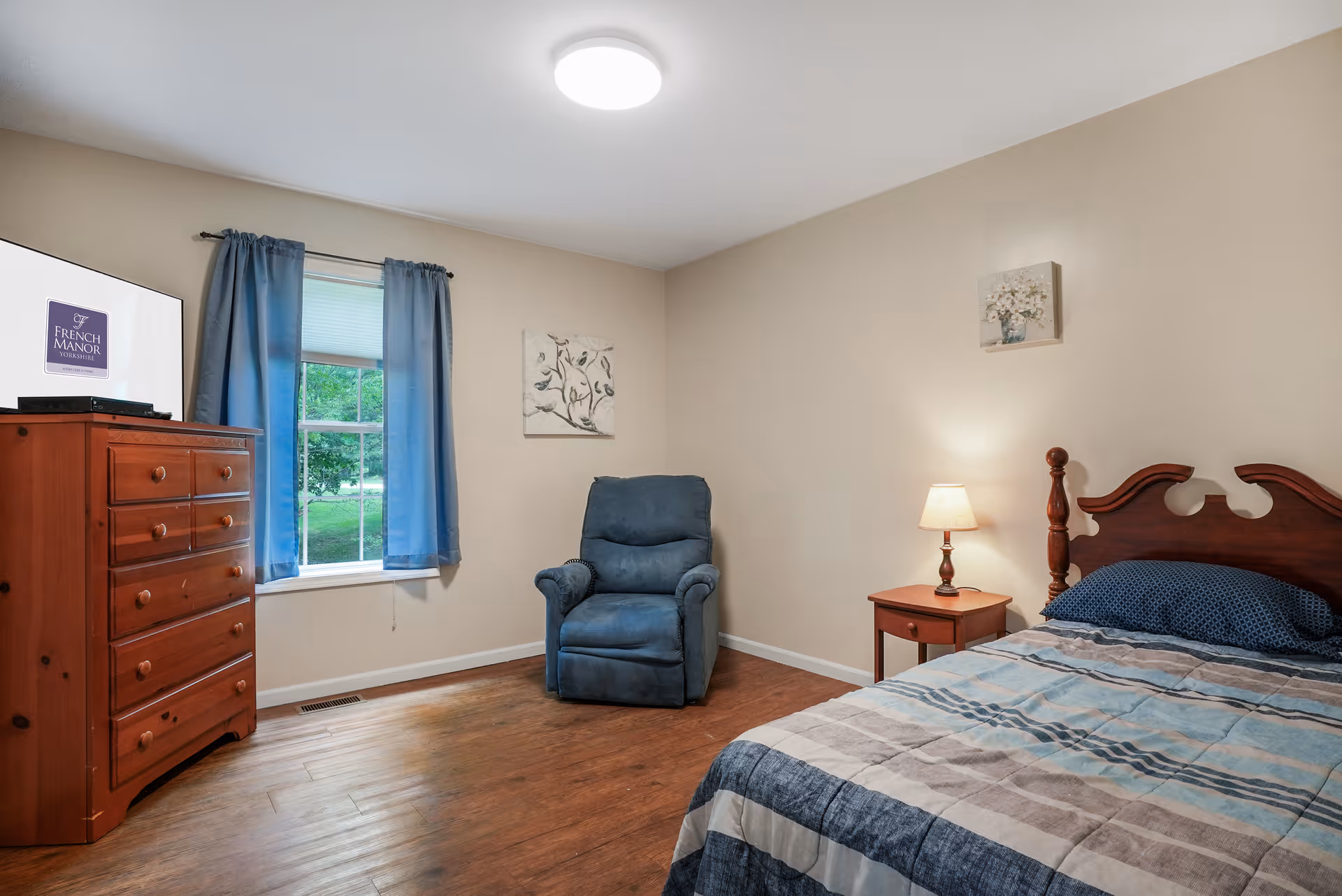 A cozy bedroom with a wooden bed featuring a blue and gray striped comforter, a wooden nightstand with a lit lamp, a blue recliner chair, a wooden dresser, and a window with blue curtains. The walls are beige and decorated with two small paintings.