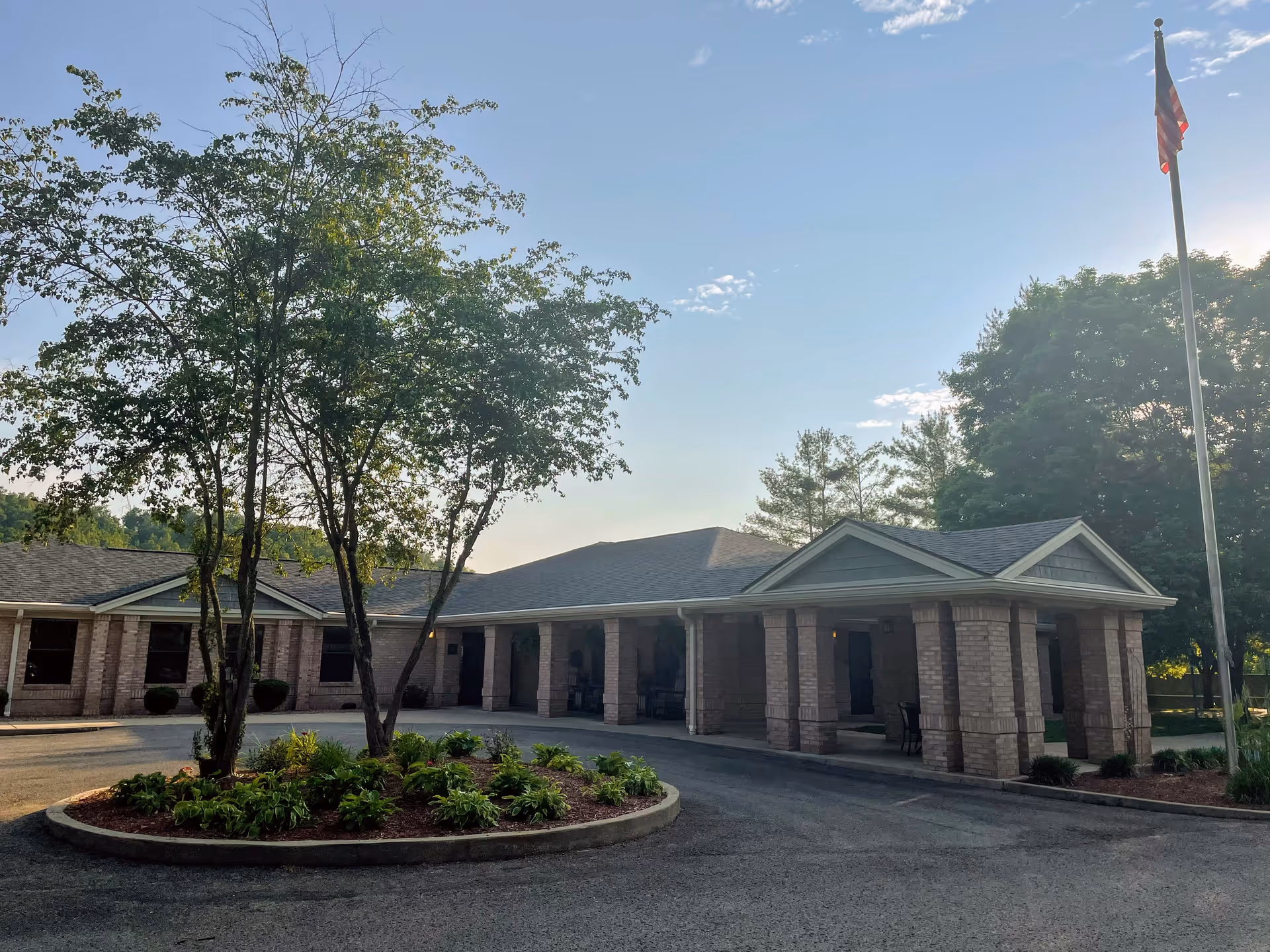 Exterior view of Elliott Nursing and Rehabilitation facility showing a single-story brick building with a covered entrance supported by columns. There is a circular landscaped area with trees and plants in front, and an American flag on a tall flagpole to the right. The sky is clear with some clouds and the sun is low, casting shadows.