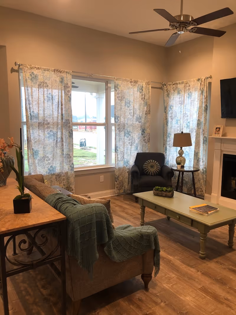 A cozy living room with a beige sofa draped with a green throw blanket, a green coffee table with a small plant and books on it, a dark armchair with a decorative pillow, a side table with a lamp, floral curtains on large windows, a ceiling fan, and a fireplace with a mounted TV above it.