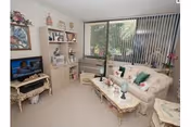 A cozy living room with a cream-colored sofa adorned with green and pink cushions, a glass coffee table with decorative items, a TV on a small stand, and a shelving unit with various knick-knacks. Large vertical blinds cover the window, allowing natural light to enter the room.