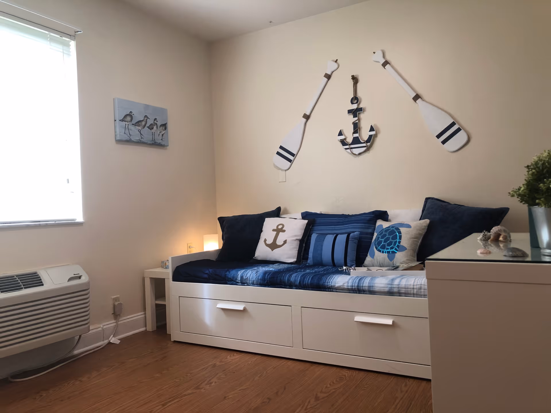 A cozy interior room with a white daybed featuring blue and white cushions, including nautical-themed pillows with an anchor and a turtle. Above the daybed, two white oars and a striped anchor decoration hang on the wall. There is a small white side table with a lamp next to the daybed, a window with blinds on the left, and a white air conditioning unit below the window. A small plant and seashell decorations are on a white cabinet to the right.