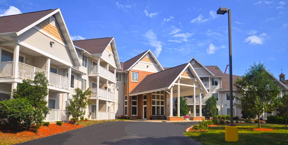 Exterior view of a senior living facility named Meadowmere & Mitchell Manor West Allis, showing a multi-story building with balconies, a covered entrance, landscaped greenery, and a clear blue sky.