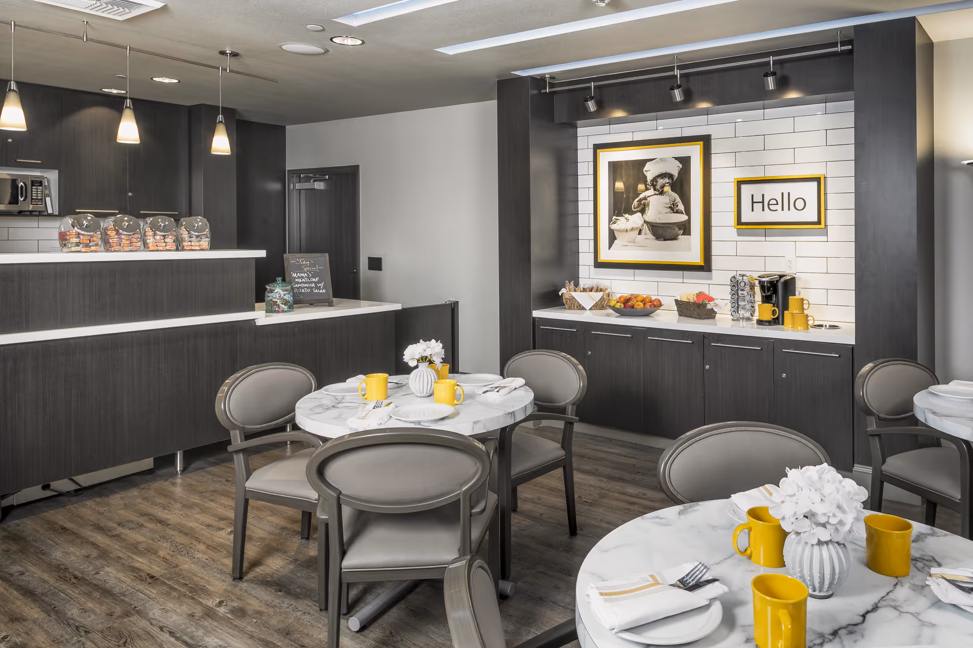 Modern communal dining area with round marble tables, gray chairs, a serving counter and a framed 'Hello' sign on a tiled wall.