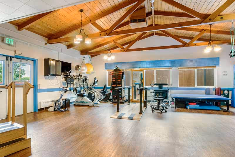 A spacious rehabilitation or physical therapy room with wooden floors and a high wooden ceiling with exposed beams. The room contains exercise equipment including stationary bikes, parallel bars for walking practice, therapy tables, and various rehabilitation tools. Large windows with blinds allow natural light to enter, and there is an exit door on the left side.