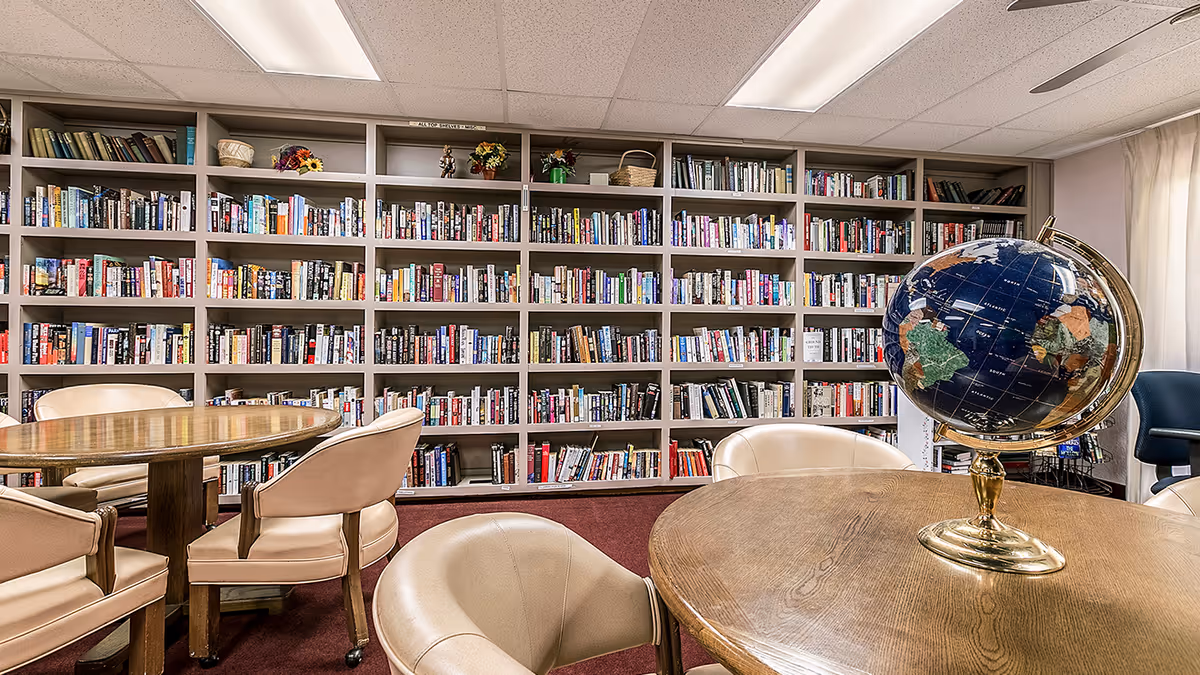 A cozy library room with multiple wooden tables and beige chairs arranged around them. A large bookshelf filled with numerous books covers the entire back wall. A decorative globe sits on one of the tables. The room has a carpeted floor and fluorescent ceiling lights.
