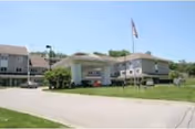 Exterior view of a two-story senior living facility with a covered entrance, an American flag on a flagpole, and green lawn surrounding the building under a clear sky.