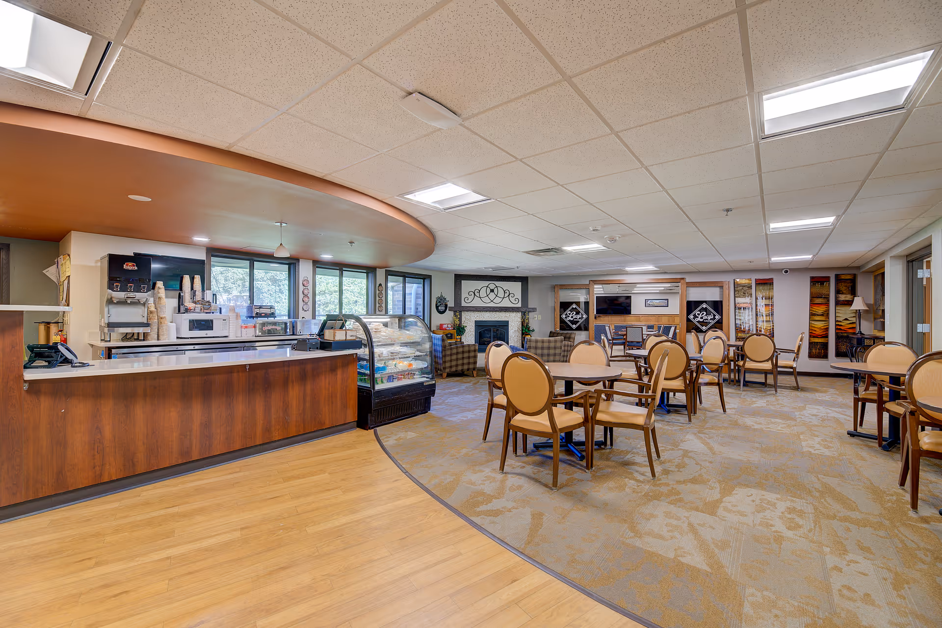 Open communal dining and café area with a service counter and refrigerated display case on the left and round dining tables and chairs across the carpeted room.