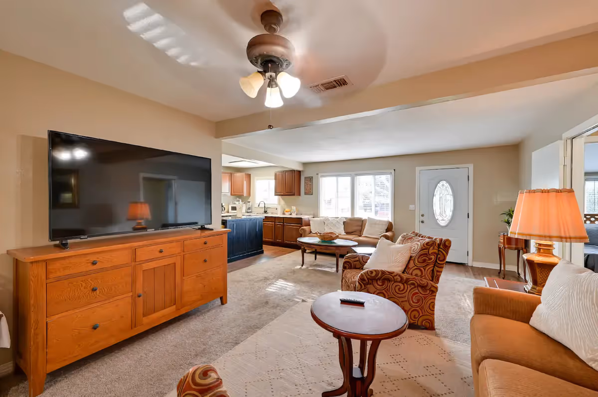 Open living room with a large flat-screen TV on a wooden dresser, multiple sofas and armchairs, coffee tables, and a view into the kitchen and front door.