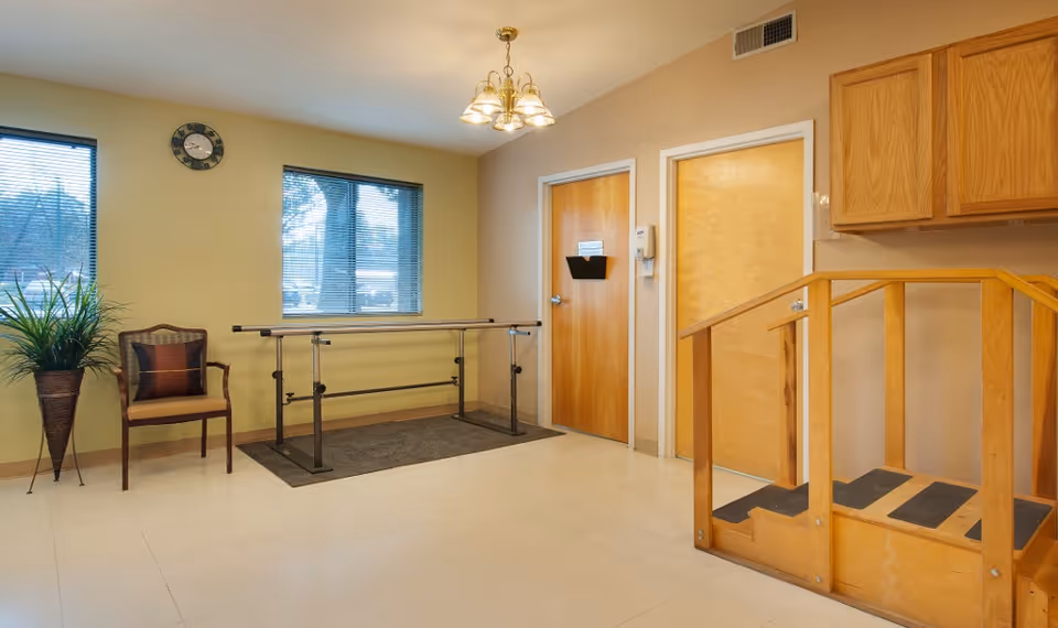 Rehabilitation therapy room featuring parallel bars, a small wooden practice staircase, a chair and potted plant under windows.