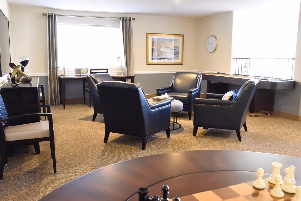 A cozy living room with four black leather armchairs arranged around a small round ottoman and table. There is a large window with curtains, a desk with a chair, a framed picture on the wall, a clock, and a shuffleboard table near the window. In the foreground, part of a chessboard with chess pieces is visible.