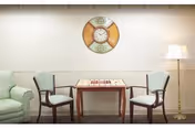 A cozy interior room with a small wooden table set for a board game, flanked by two wooden chairs with light blue cushions. To the left is a light green upholstered armchair, and to the right stands a tall floor lamp with a white lampshade. A decorative wall clock with a circular design hangs on the beige wall above the table.