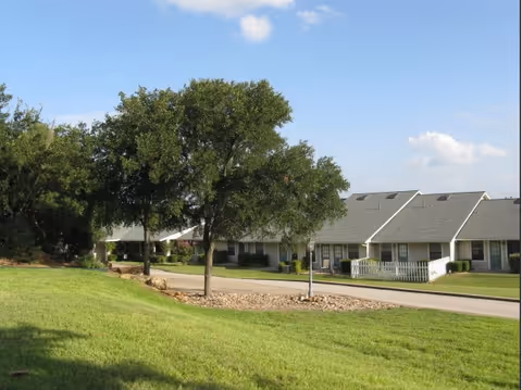 View of a residential area in Marble Falls Assisted Living with green grass, a tree in the foreground, and single-story buildings with gray roofs and white fences under a blue sky with some clouds.