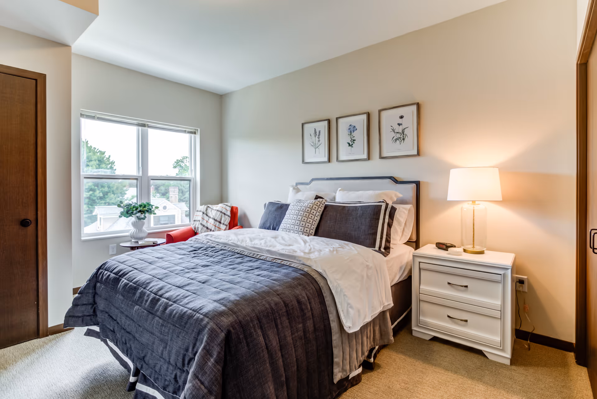 Well-lit bedroom with a neatly made bed, nightstand and lamp, framed botanical prints, and a window with a red chair.