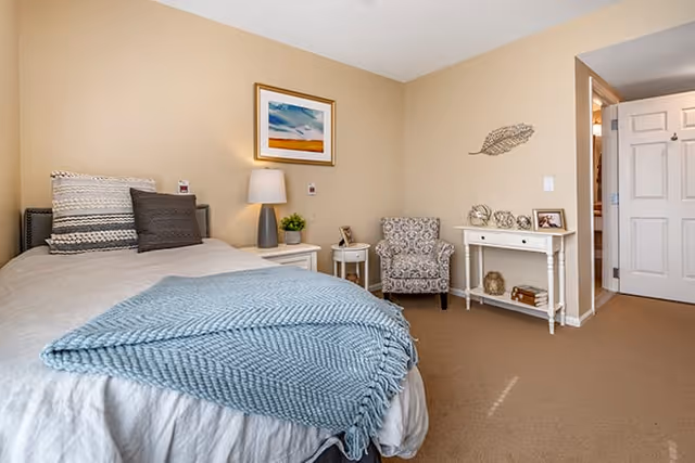 A cozy bedroom with a neatly made bed featuring a light blue knitted blanket and multiple pillows. Next to the bed is a white nightstand with a lamp and a small plant. Across from the bed is a patterned armchair and a white console table decorated with small ornaments and framed photos. The walls are painted beige, and there is a framed landscape painting above the nightstand. An open door leads to another room.
