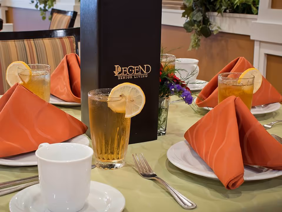 A dining table set with white plates, orange folded napkins, glasses of iced tea garnished with lemon slices, a white coffee cup, silverware, and a black menu with the text 'Legend Senior Living'. There is a small floral arrangement in the background and striped upholstered chairs.