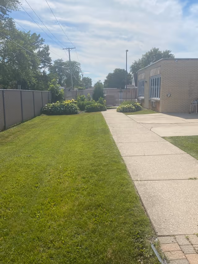 A paved walkway runs alongside a green lawn with bushes and trees, leading to a brick building with several windows. The sky is partly cloudy and there is a chain-link fence on the left side of the walkway.