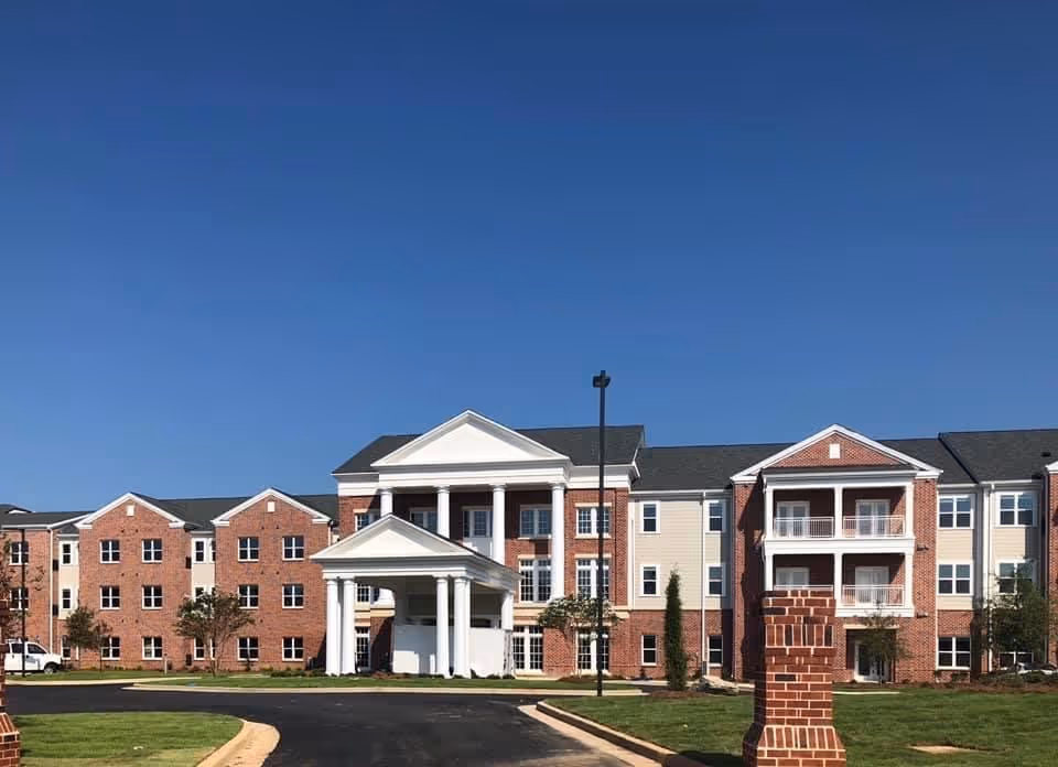 Exterior front view of a large three-story brick senior living building with a columned entrance and balconies under a clear blue sky.