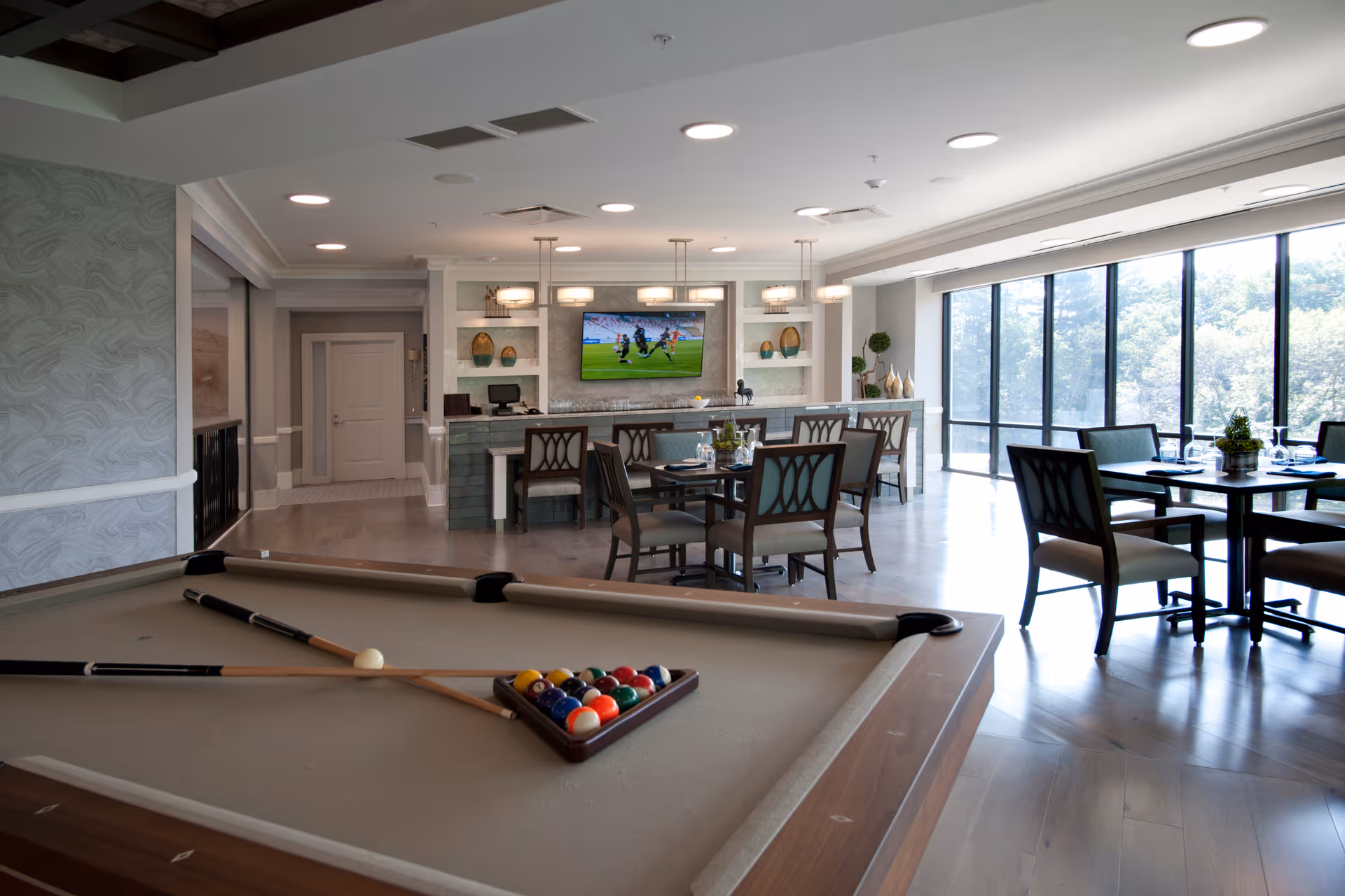 A spacious and well-lit common area in a senior living facility featuring a pool table with cues and balls arranged for a game in the foreground. In the background, there are dining tables and chairs set near large windows that let in natural light, and a bar area with stools and a wall-mounted TV showing a sports game.