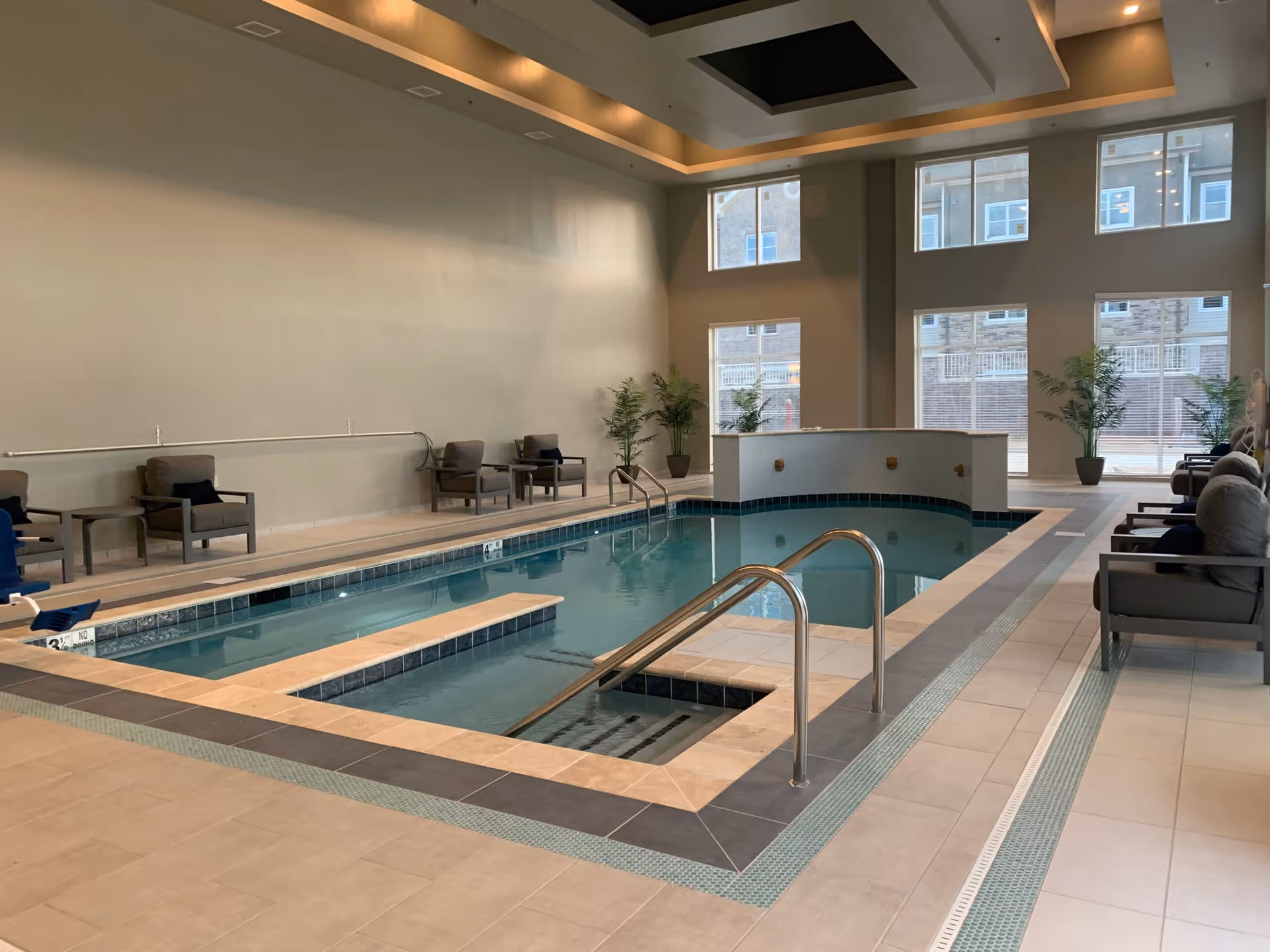 Indoor swimming pool area with a small pool and a hot tub section, surrounded by tiled flooring. Several cushioned chairs and small tables are arranged along the walls, with potted plants near large windows letting in natural light.