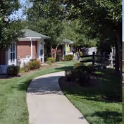 A paved walkway curves through a grassy area with small brick cottages on the left and trees providing shade on the right.