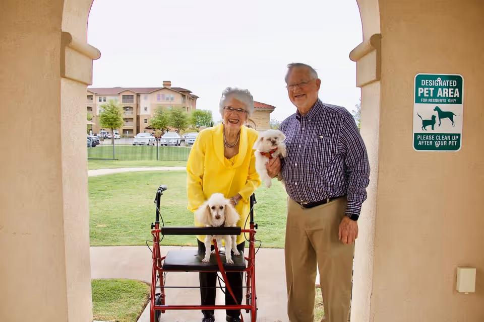 An elderly woman with a walker and an elderly man standing under an archway in an outdoor designated pet area. The woman is wearing a bright yellow top and holding a small white dog on her walker, while the man is wearing a checkered shirt and holding another small white dog. Behind them is a grassy area with trees and a building in the background.