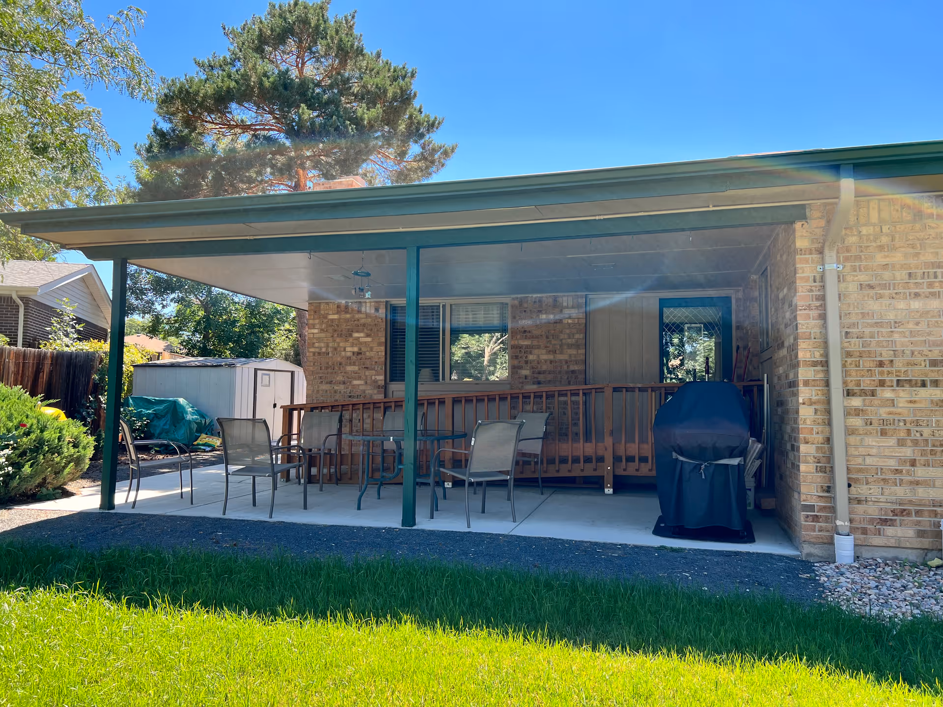 Covered patio area attached to a brick building with outdoor chairs, a table, a barbecue grill covered with a black cover, and a wooden ramp. There is green grass in the foreground and trees in the background under a clear blue sky.