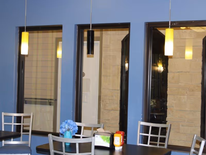 Interior view of a dining area with blue walls and three large windows. There are three pendant lights hanging from the ceiling, two with yellow shades and one with a black shade. A table with silver chairs is visible, featuring a small blue vase with blue flowers, a napkin holder, and ketchup and mustard bottles.
