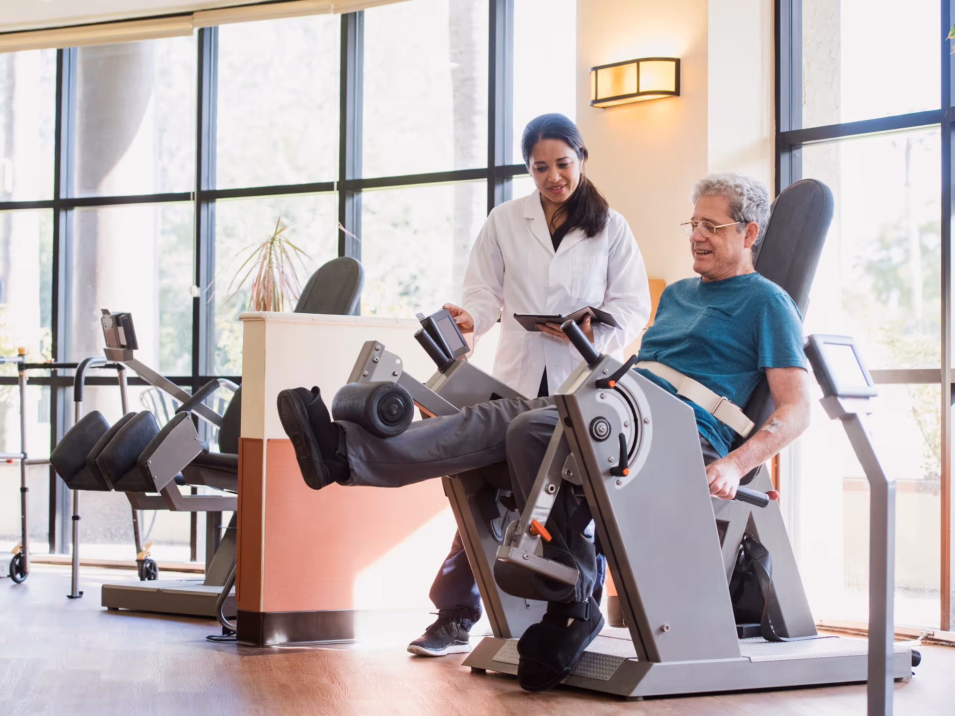 An elderly man exercises his legs on a leg press machine in a well-lit gym area with large windows. A female healthcare professional in a white coat stands beside him, holding a tablet and providing assistance.