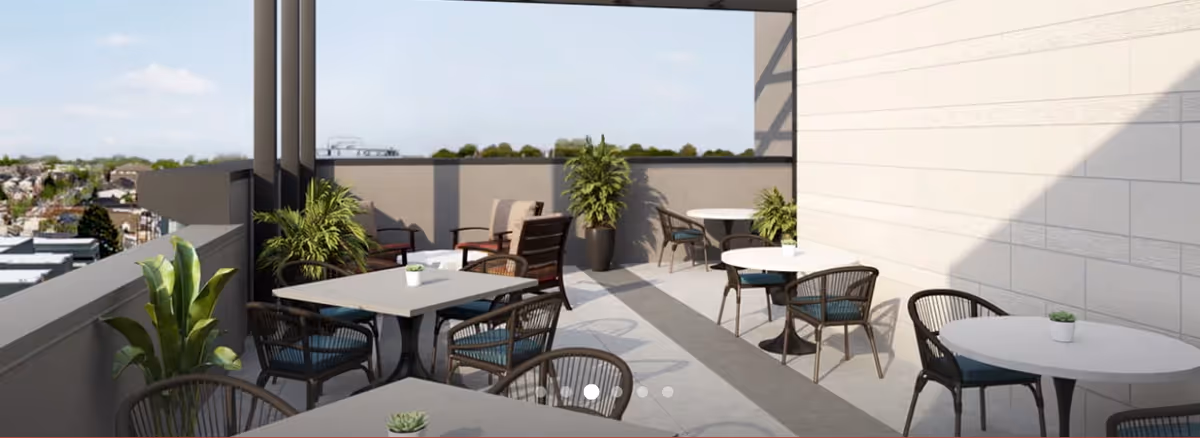 Outdoor patio area with several round and square tables surrounded by chairs with blue cushions. There are potted plants placed along the walls and on the tables. The patio overlooks a distant view of buildings and greenery under a clear sky.