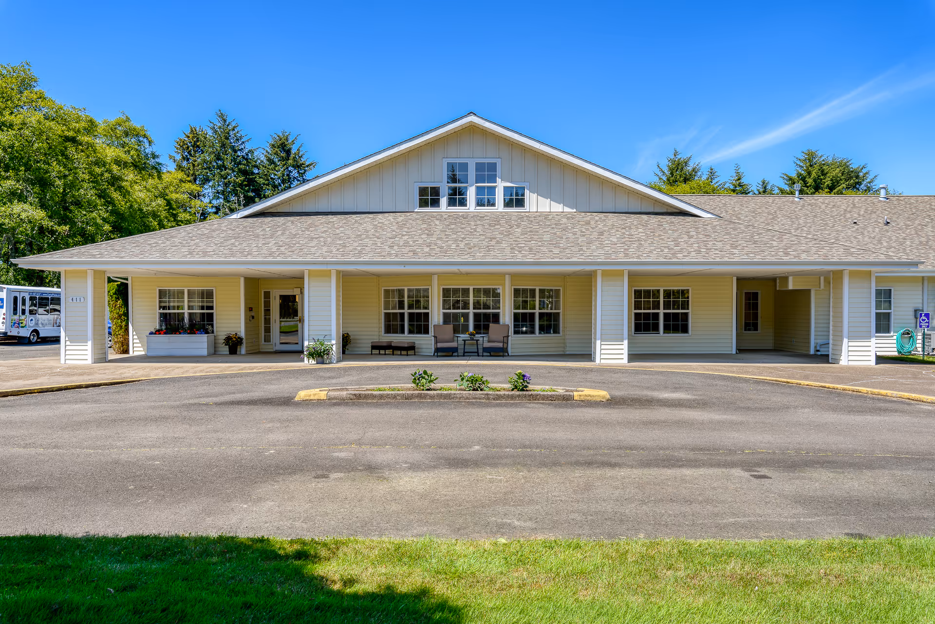Front exterior view of a single-story building with a covered entrance, beige siding, multiple windows, and outdoor seating. There is a circular driveway with a small landscaped island in front, green grass in the foreground, and trees in the background under a clear blue sky.