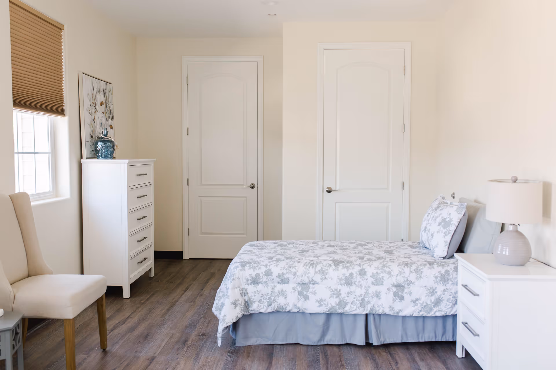 A bright and clean bedroom with a single bed covered in a floral patterned bedspread and matching pillow. The room features two white doors, a white chest of drawers with a decorative vase and framed artwork on top, a beige upholstered chair, a white nightstand with a lamp, and a window with a brown blind. The floor is wooden and the walls are painted light beige.