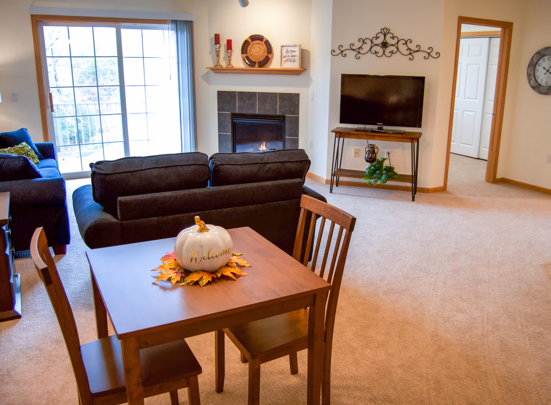A cozy living room with a small wooden dining table and two chairs in the foreground. The table has a white decorative pumpkin with the word 'Welcome' on it, surrounded by autumn leaves. Behind the table are two dark-colored sofas facing a fireplace with a lit fire. Above the fireplace is a wooden shelf with decorative items. To the right, a flat-screen TV is mounted on a wooden stand with a plant underneath. A sliding glass door lets in natural light from outside.