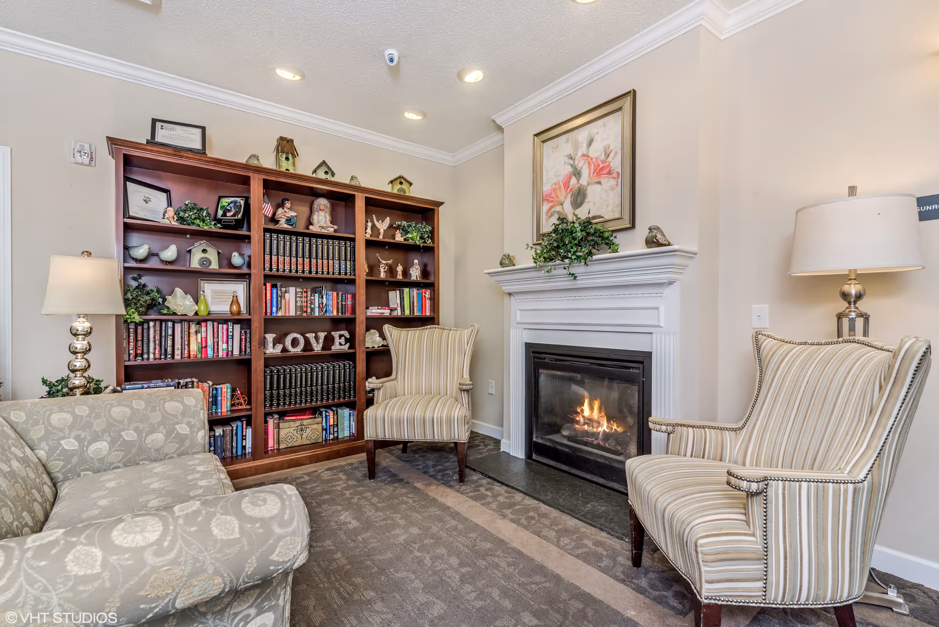 A cozy living room area with a lit fireplace, two striped armchairs, a patterned sofa, a wooden bookshelf filled with books and decorative items, and two table lamps on either side of the room. A floral painting hangs above the fireplace.