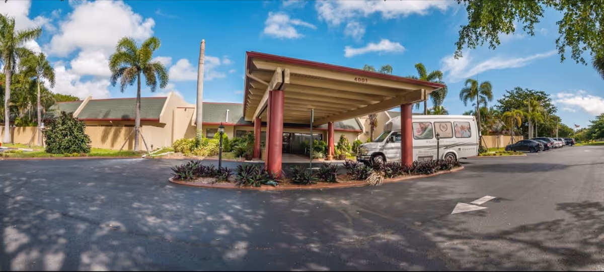 Exterior view of Willow Bay Senior Resort showing the entrance with a covered drop-off area supported by red columns. A white shuttle van with the facility's branding is parked near the entrance. The building has a beige facade with a green roof, surrounded by palm trees and landscaping. Several cars are parked along the driveway under a partly cloudy blue sky.