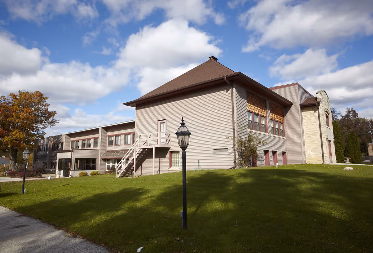 Two-story beige building with exterior stairs and lamp posts on a green lawn under a partly cloudy sky.