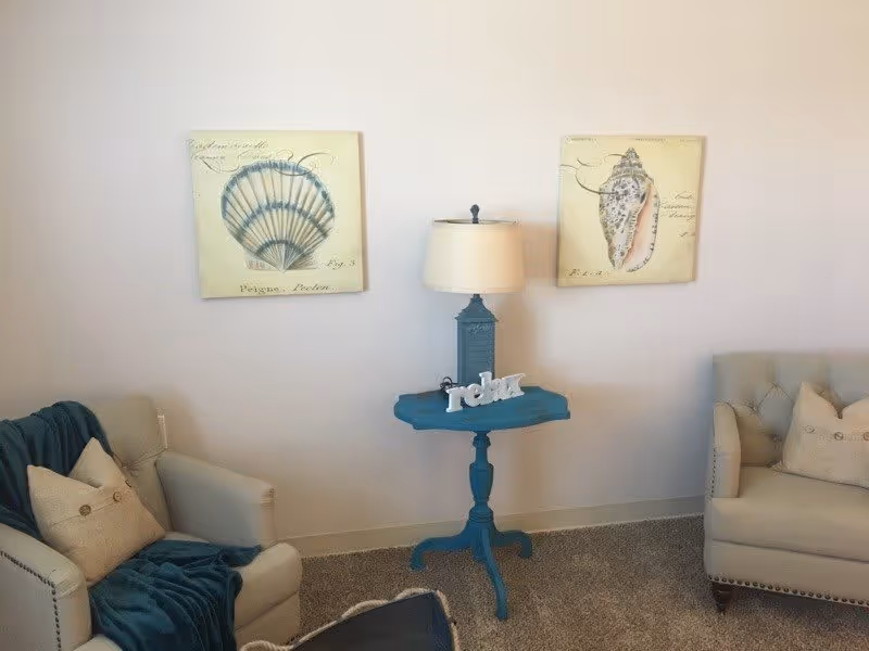 A cozy sitting area with two beige armchairs, each with a pillow. A blue throw blanket is draped over the left armchair. Between the chairs is a small blue side table with a lamp and a decorative 'relax' sign. Two framed pictures of seashells hang on the wall behind the table.