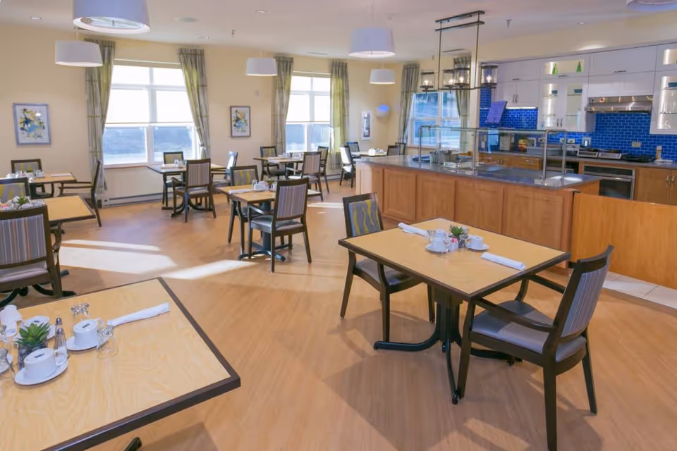 A bright and clean dining room with multiple tables and chairs arranged neatly. The tables are set with white napkins, cups, and small plants. Large windows with green curtains allow natural light to fill the room. The back of the room features a kitchen area with white cabinets, a blue tiled backsplash, and stainless steel appliances.