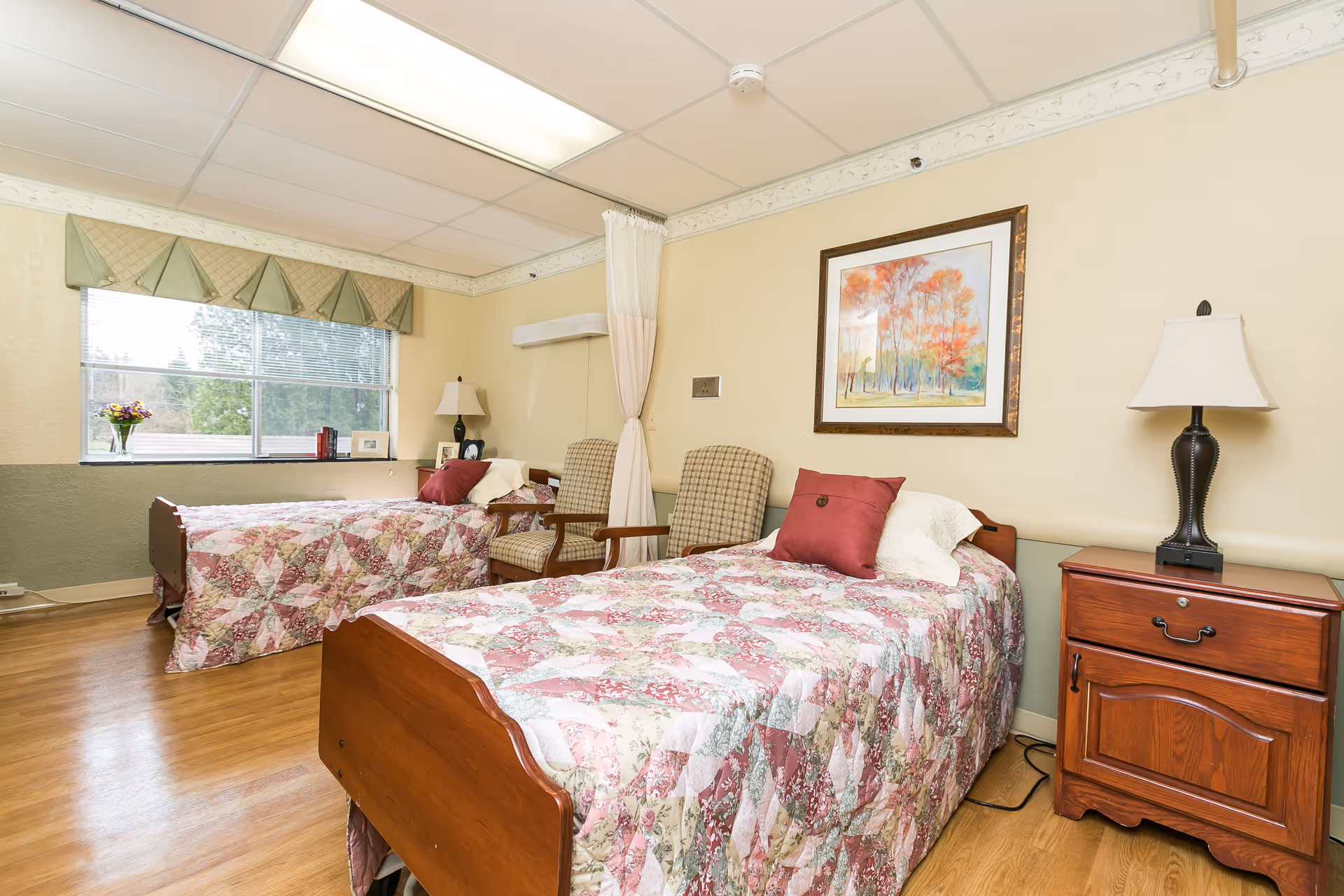 Bright nursing-facility bedroom with two twin beds covered in floral quilts, wooden nightstands, chairs, and a large window.